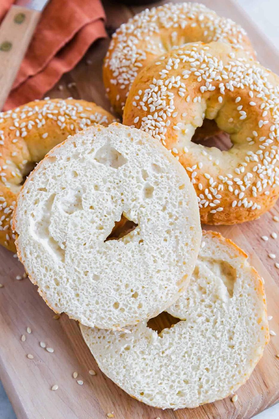 A close-up of five sesame seed high protein bagels on a wooden surface. One bagel is sliced in half, showing its soft interior. A rust-colored cloth napkin is partially visible in the background.