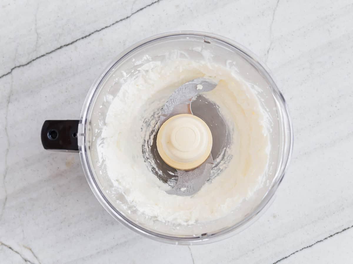Top view of a food processor with whipped cottage cheese inside, sitting on a marble countertop. The plastic and metal blade is visible, showing a smooth texture of the mixture.
