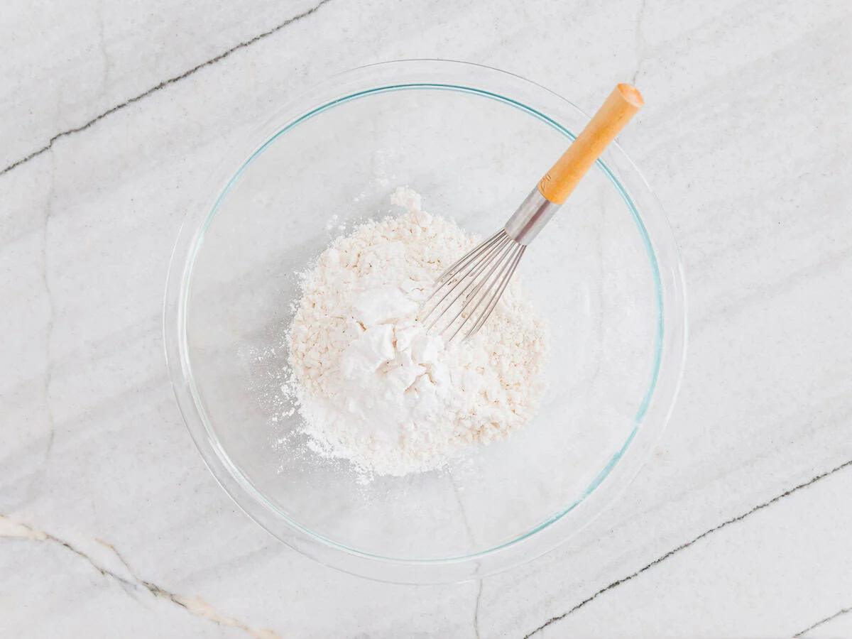 A glass bowl containing flour is placed on a light marble countertop. A whisk with a wooden handle is resting in the flour, ready for mixing.