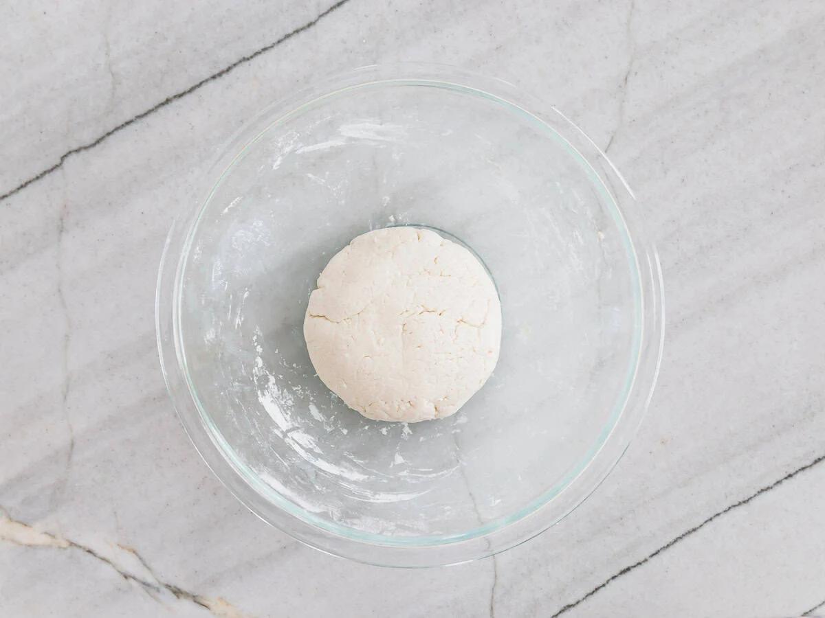 A ball of dough sits in a transparent glass bowl on a light gray, marble-textured surface. The dough is smooth and placed at the center of the bowl.