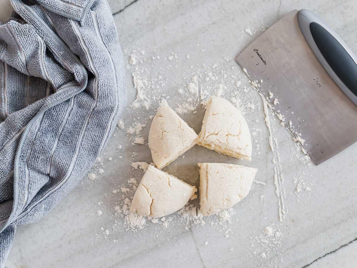 Bagel dough on a floured surface, cut into quarters. A metal dough scraper is nearby and a gray striped towel lies to the side.