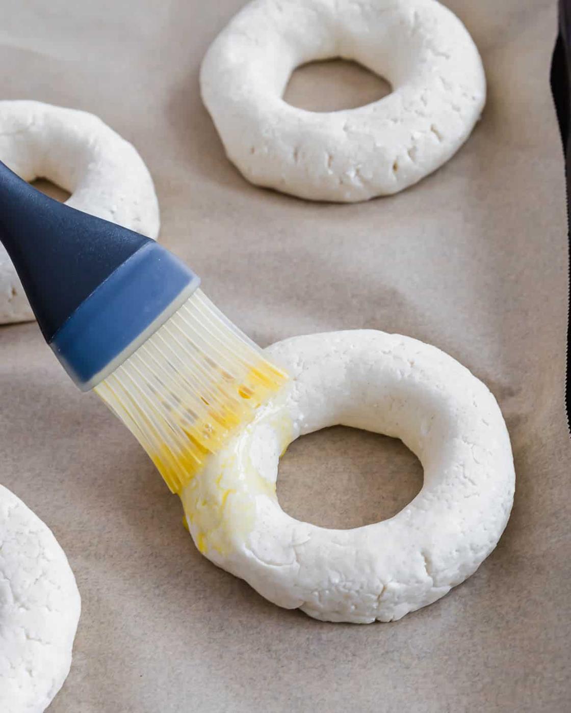 A silicone brush applies egg wash to an unbaked bagel on parchment paper. The dough is light in color and the scene suggests preparation for baking.