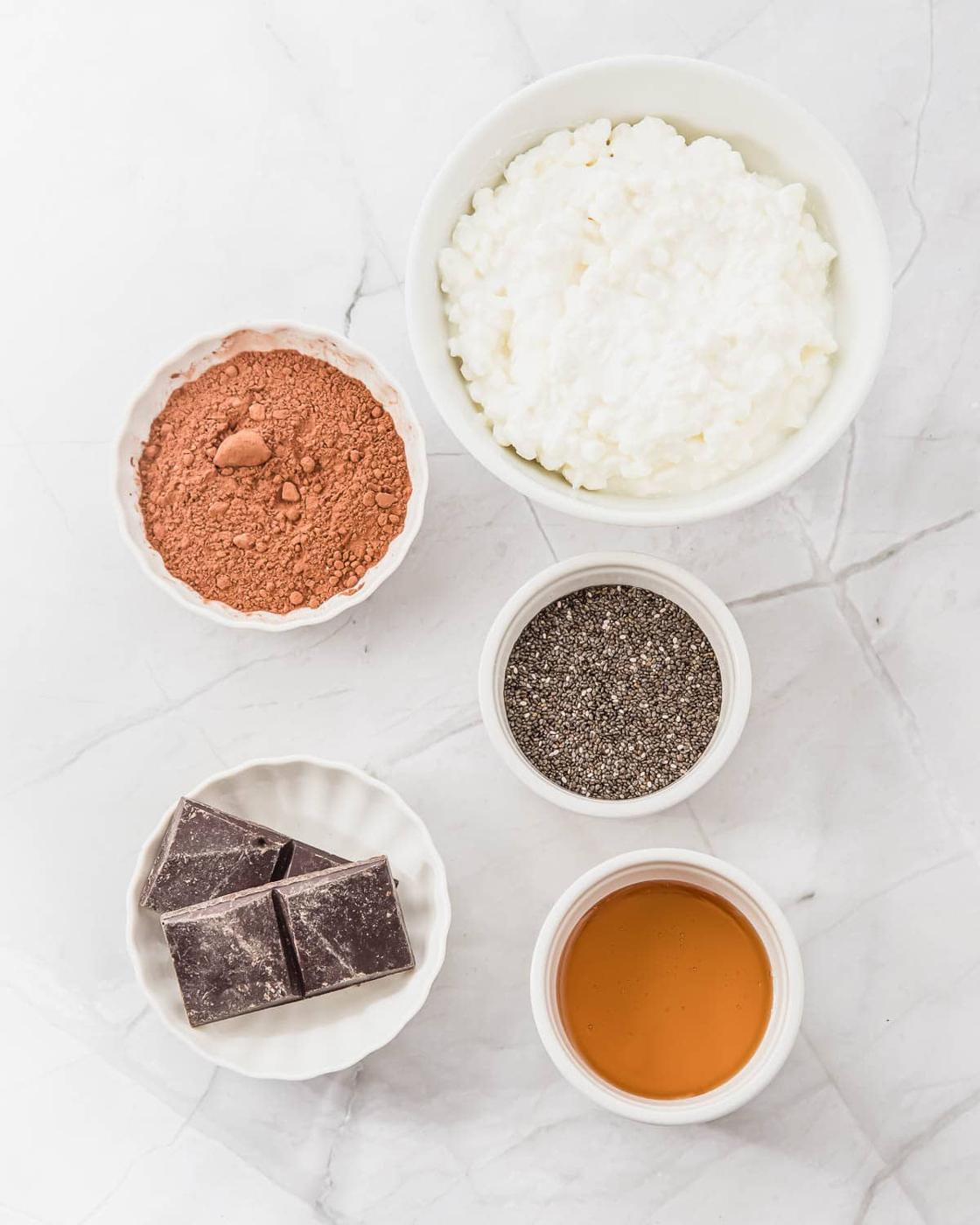 Bowls on a marble surface containing cottage cheese, cocoa powder, chia seeds, chocolate chunks, and honey.