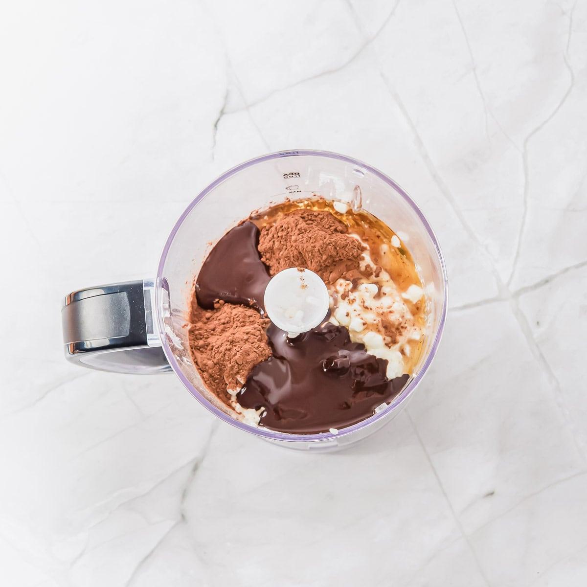 A food processor bowl on a marble countertop containing chocolate sauce, cocoa powder, and cottage cheese, ready to be blended.