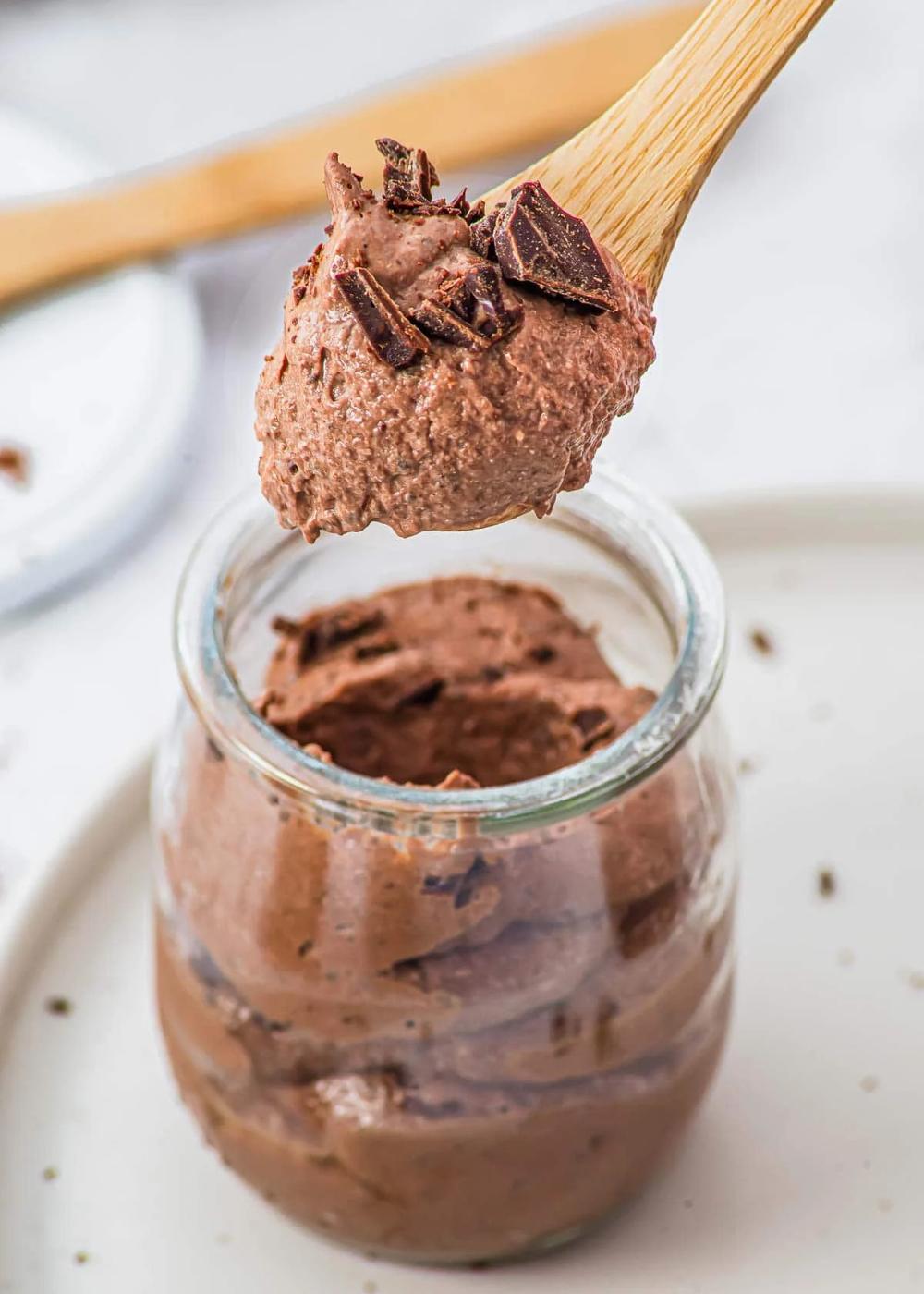 A jar filled with high protein chocolate mousse topped with chocolate shavings. A spoon lifts a portion of the mousse, showing its creamy texture. The scene is set on a white surface, with a wooden spoon partially visible in the background.