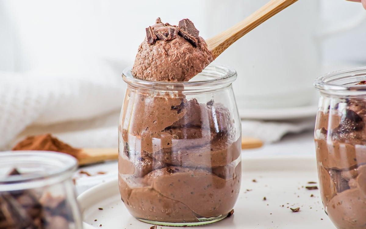 A glass jar filled with cottage cheese chocolate mousse, topped with chocolate shavings. A wooden spoon holds a scoop of the mousse. In the background are more jars and cocoa powder on a white tray and surface.