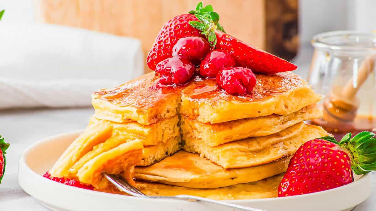 A stack of cottage cheese pancakes is topped with strawberries, raspberries, and syrup on a white plate. A fork rests on the plate, cutting a section from the stack. A jar of honey and a white napkin are in the background.