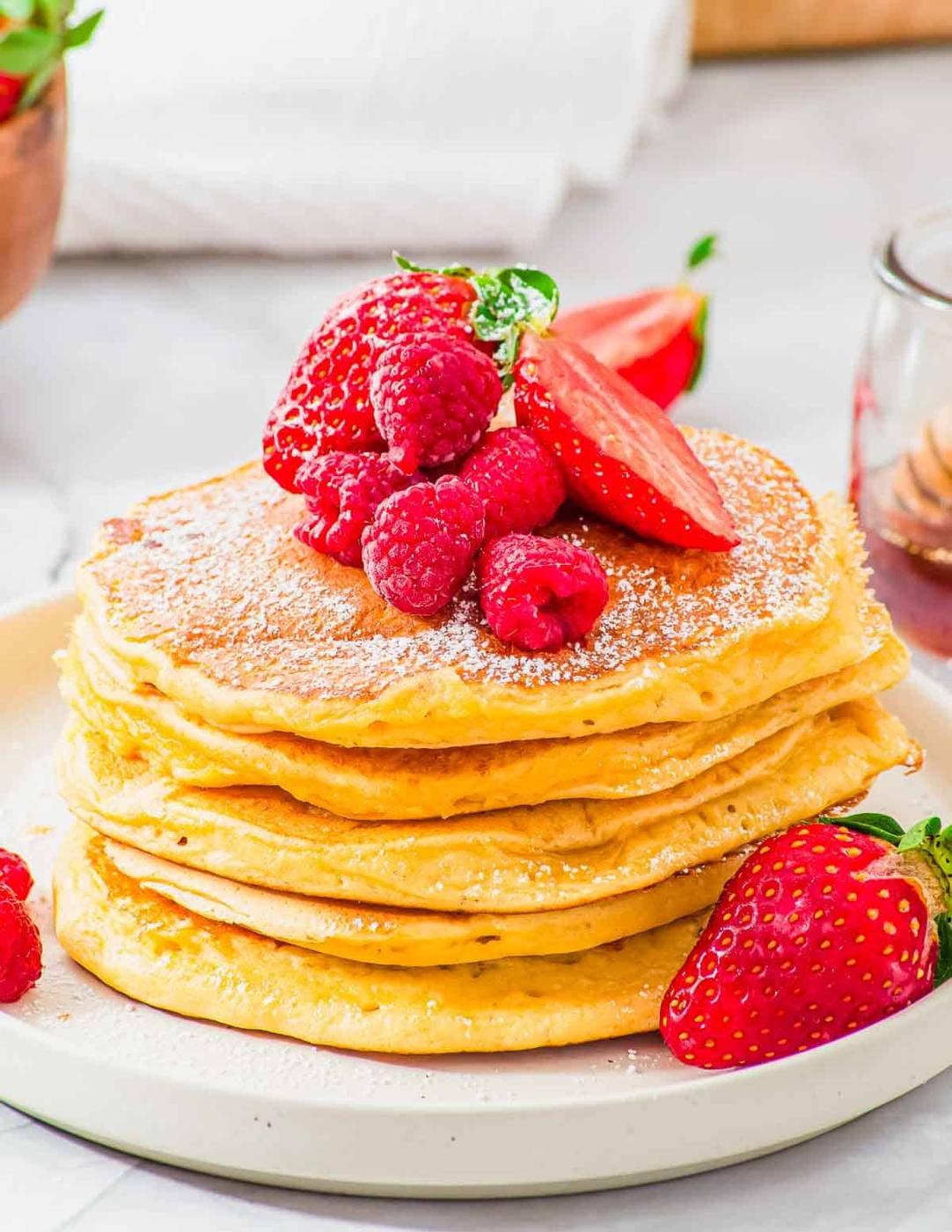A stack of golden brown cottage cheese pancakes topped with fresh raspberries, sliced strawberries, and a dusting of powdered sugar on a white plate. A jar of honey and a napkin are in the background.