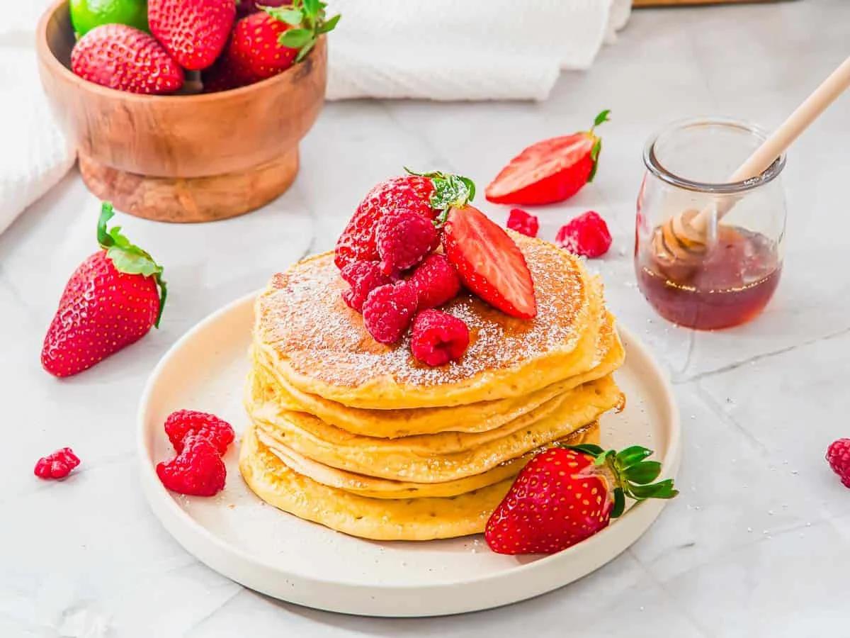 A stack of cottage cheese pancakes on a plate, topped with powdered sugar, raspberries, and a sliced strawberry. Fresh strawberries and raspberries are scattered around. A jar of honey with a dipper is beside the plate, and a bowl of strawberries is in the background.