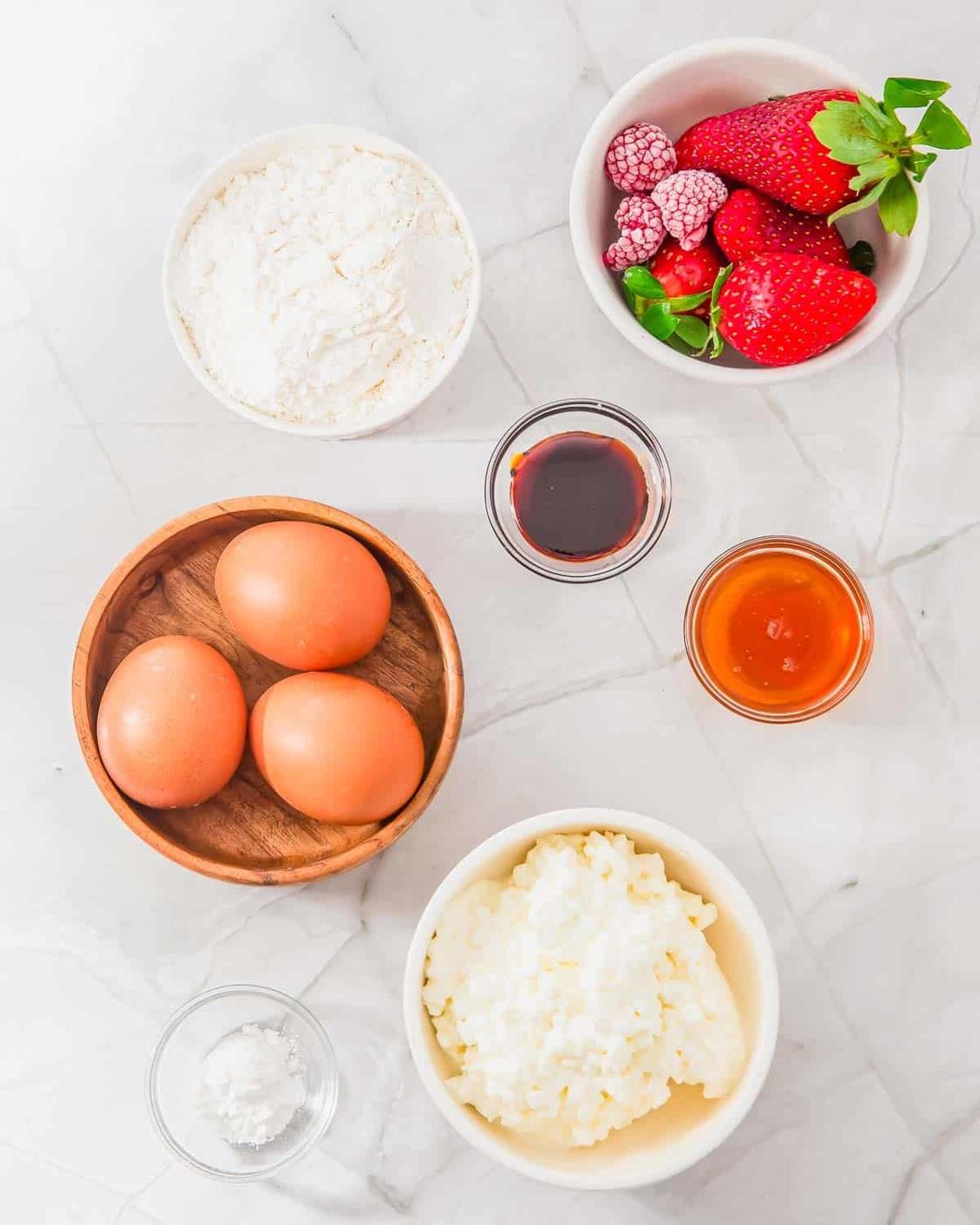 Overhead view of pancakes ingredients on a marble surface: eggs, flour, cottage cheese, honey, vanilla extract, baking soda, and a bowl of mixed berries including strawberries and raspberries.