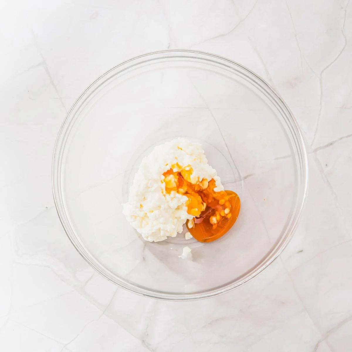 A glass mixing bowl on a marble surface contains cottage cheese and honey.