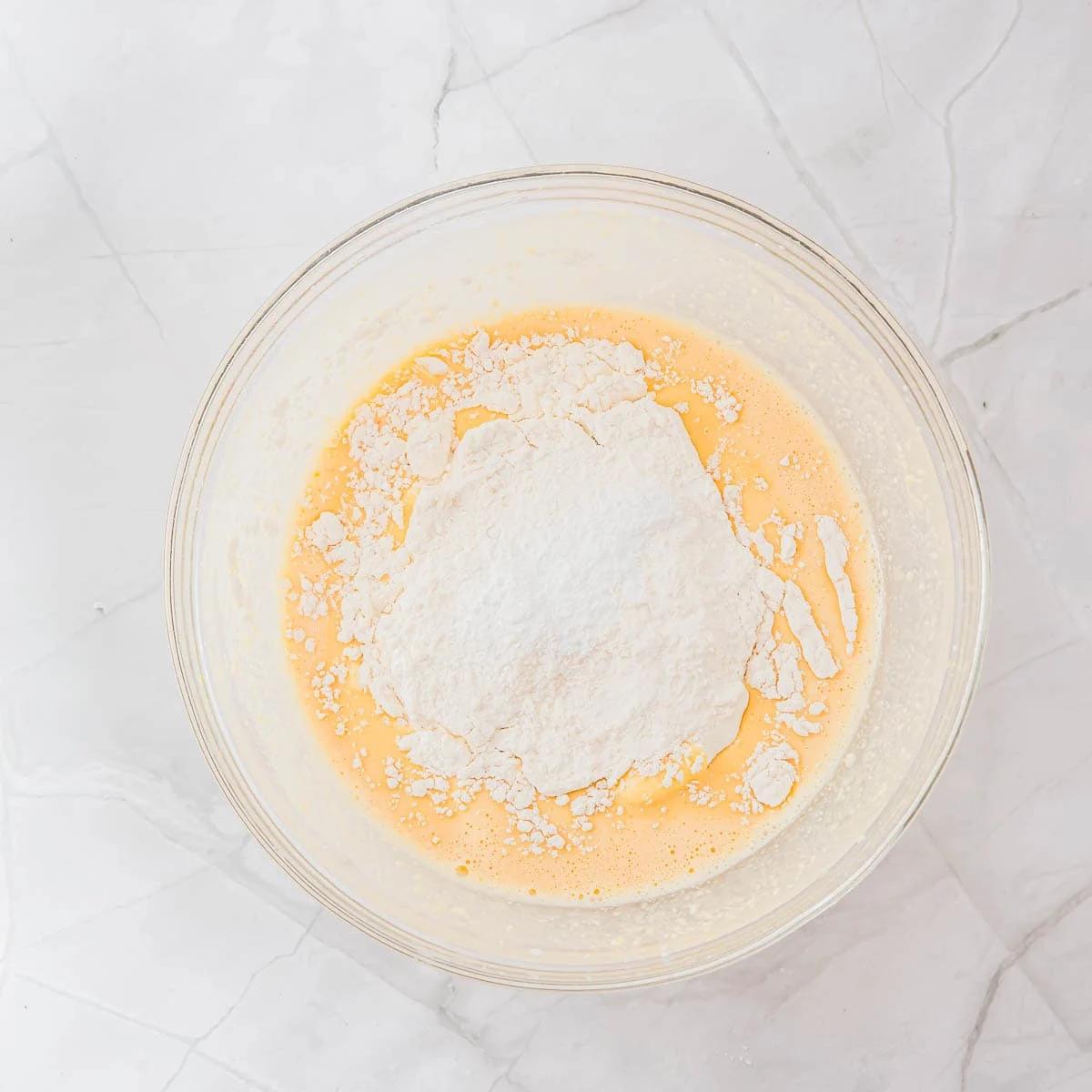 A clear glass bowl on a white marble surface contains a pancake batter with a mound of flour on top. The mixture looks ready to be stirred or blended for baking.