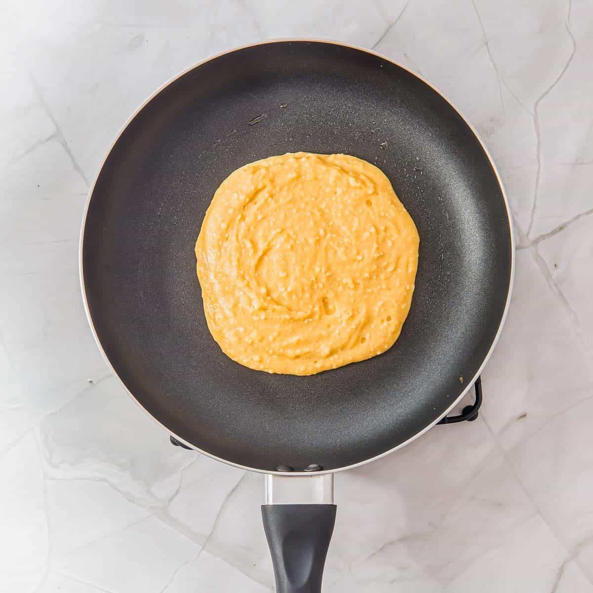 A non-stick frying pan with pancake batter in the center, resting on a marble surface.