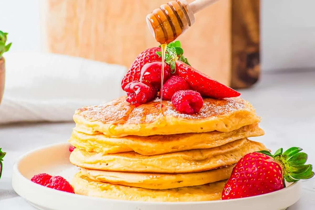 A stack of fluffy golden cottage cheese pancakes topped with fresh strawberries and raspberries on a white plate. Honey is being drizzled from a wooden dipper. A wooden board and a white cloth are in the blurred background.