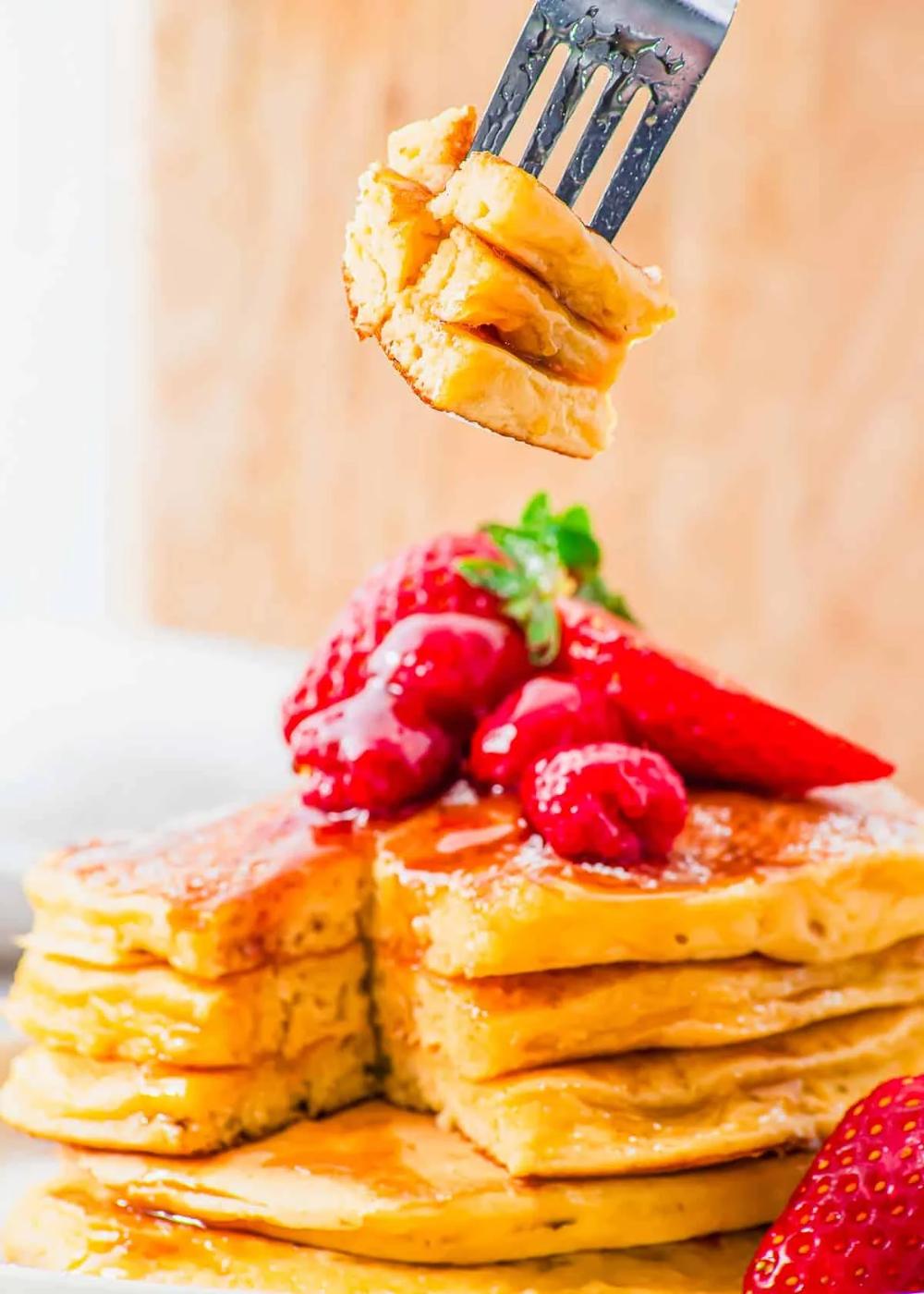 A fork holds a bite-sized piece of fluffy cottage cheese pancakes drizzled with syrup. The stack of pancakes on the plate is topped with strawberries and raspberries. The background is softly blurred, highlighting the golden texture of the pancakes.