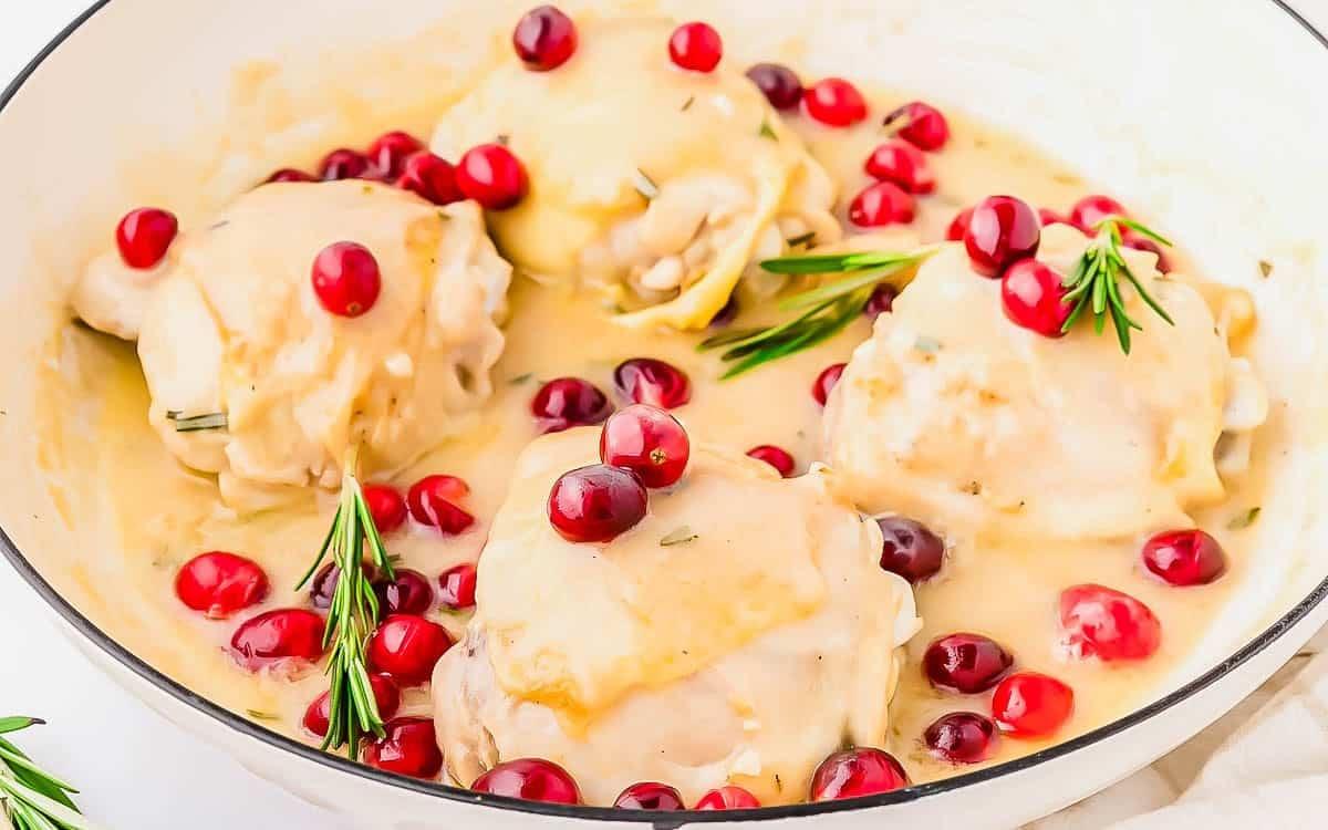 Four chicken thighs in creamy sauce are garnished with fresh cranberries and rosemary sprigs in a white dish. The sauce is thick and glossy, surrounding the chicken and vibrant cranberries.