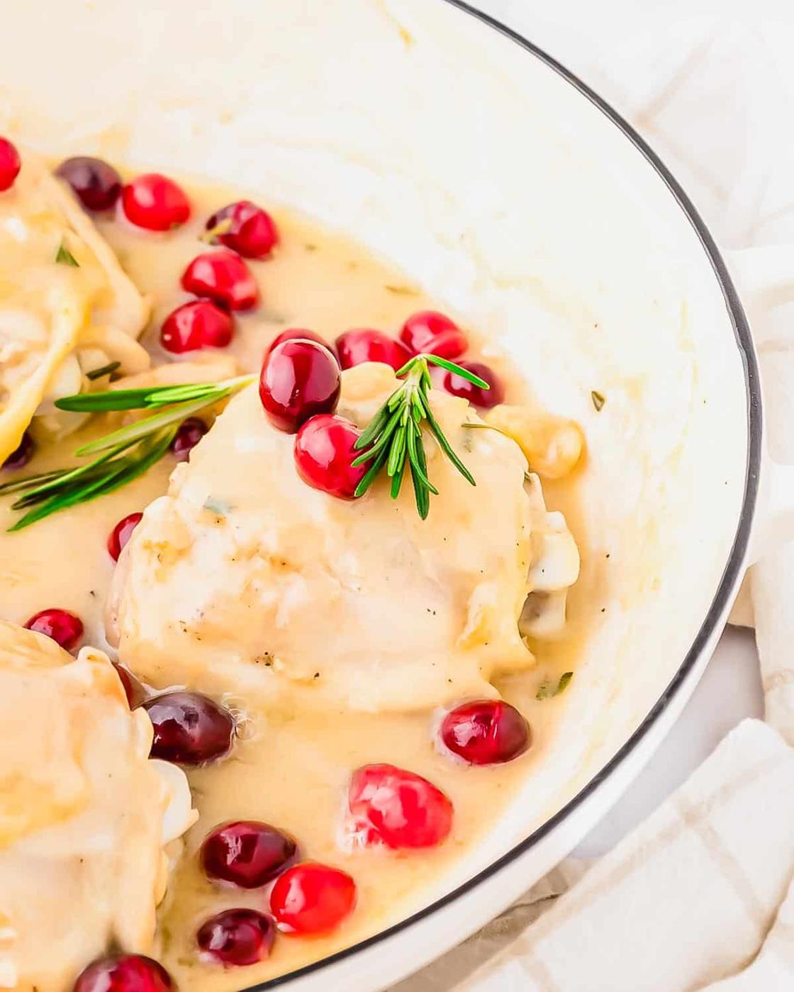 Chicken breasts in creamy white sauce, garnished with fresh cranberries and rosemary, served in a white dish with a beige-striped cloth nearby.