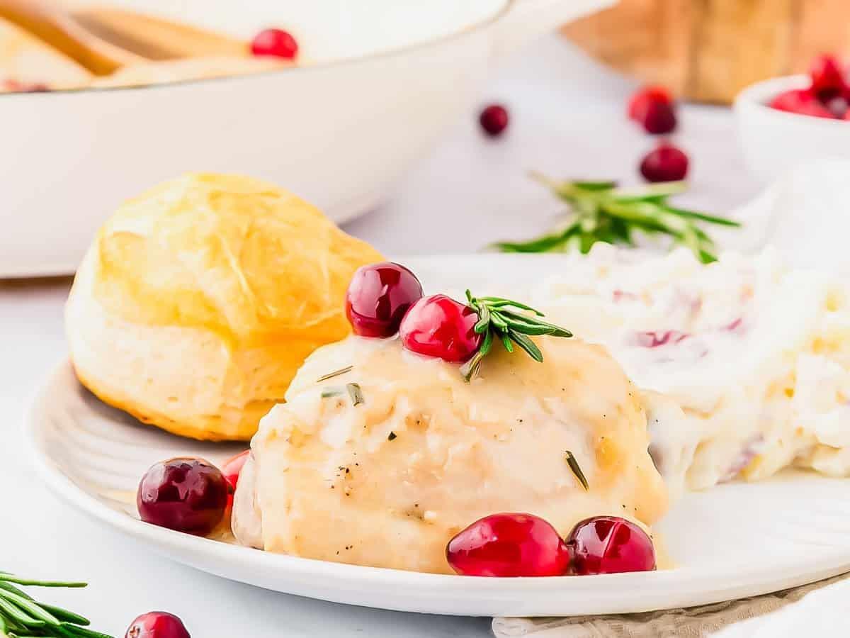 A plate with creamy cranberry chicken topped with fresh rosemary and served with mashed potatoes and a biscuit. Fresh cranberries and herbs are scattered around, creating a festive, holiday meal look.