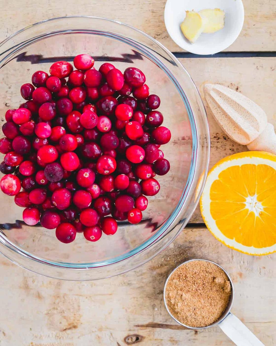 Fresh cranberries in a glass bowl with half an orange, fresh ginger slices and coconut sugar on a wooden surface.