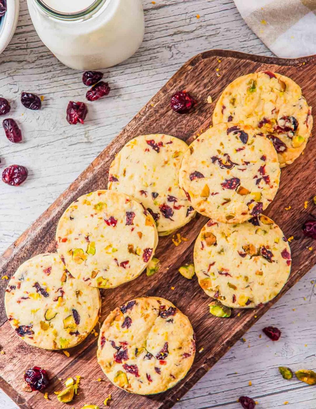 A wooden board displaying several cranberry orange pistachio shortbread cookies. The cookies are golden with visible chunks of cranberries and pistachios. Dried cranberries are scattered around. A jar of milk is partially visible in the background.