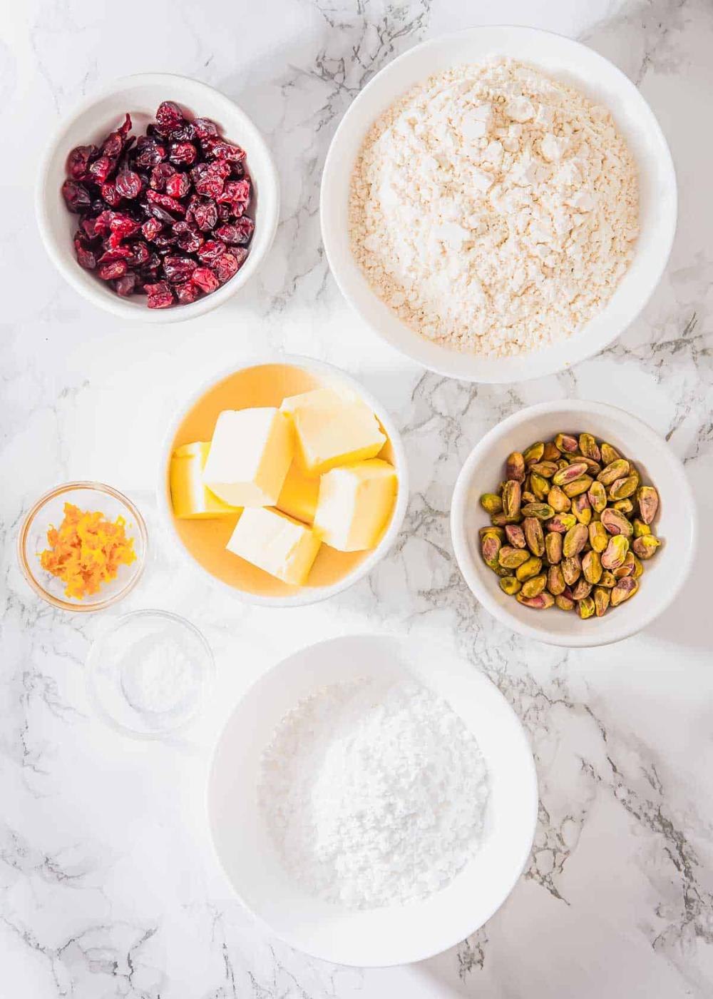 An overhead shot of baking ingredients on a marble surface. Displayed are bowls containing dried cranberries, flour, chopped pistachios, and powdered sugar. Includes a dish with butter cubes, orange zest in a small bowl, and a bit of salt.