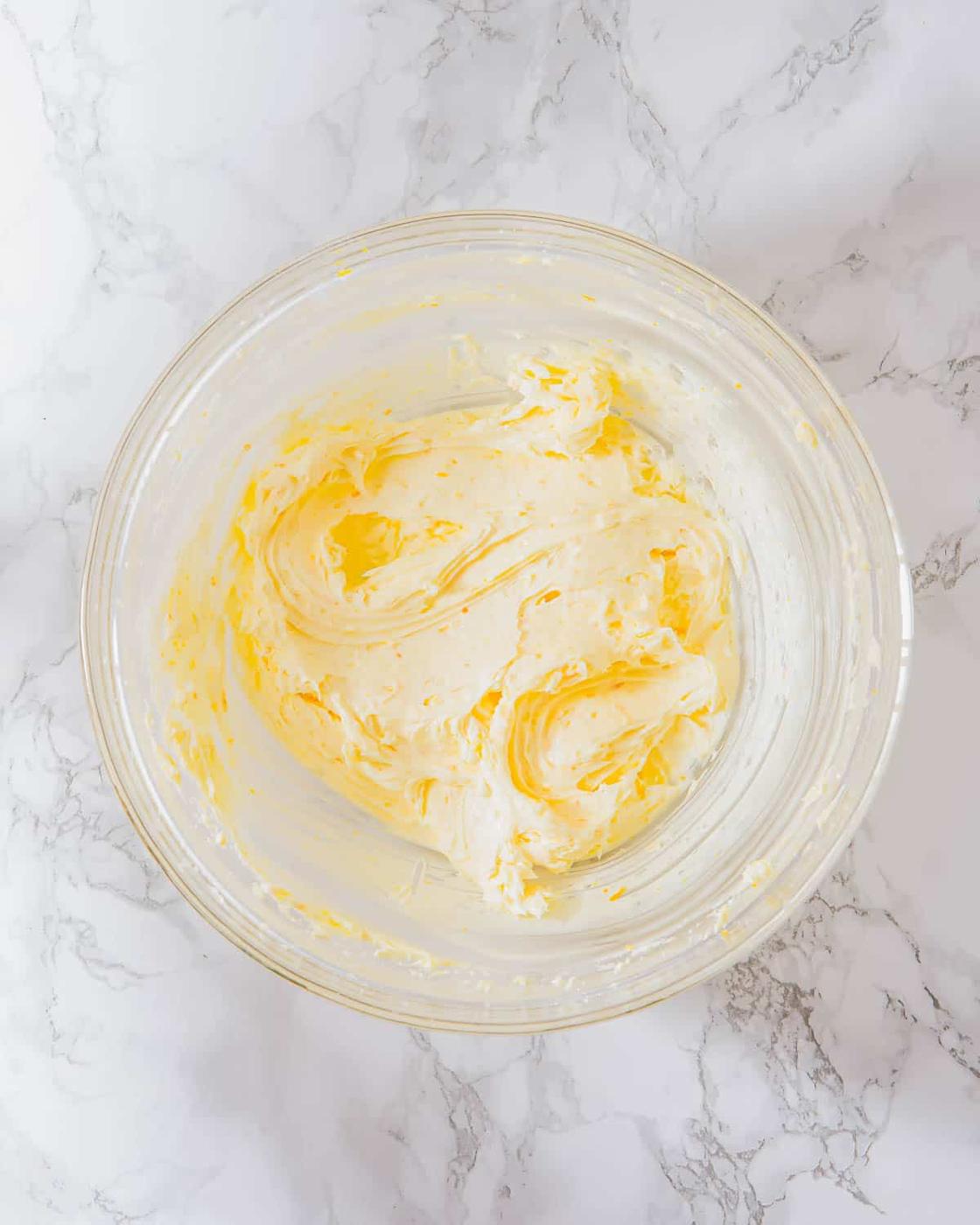 A glass bowl filled with creamed butter and sugar sits atop a white marble surface. The butter is smooth and pale yellow, with swirls visible in its texture.