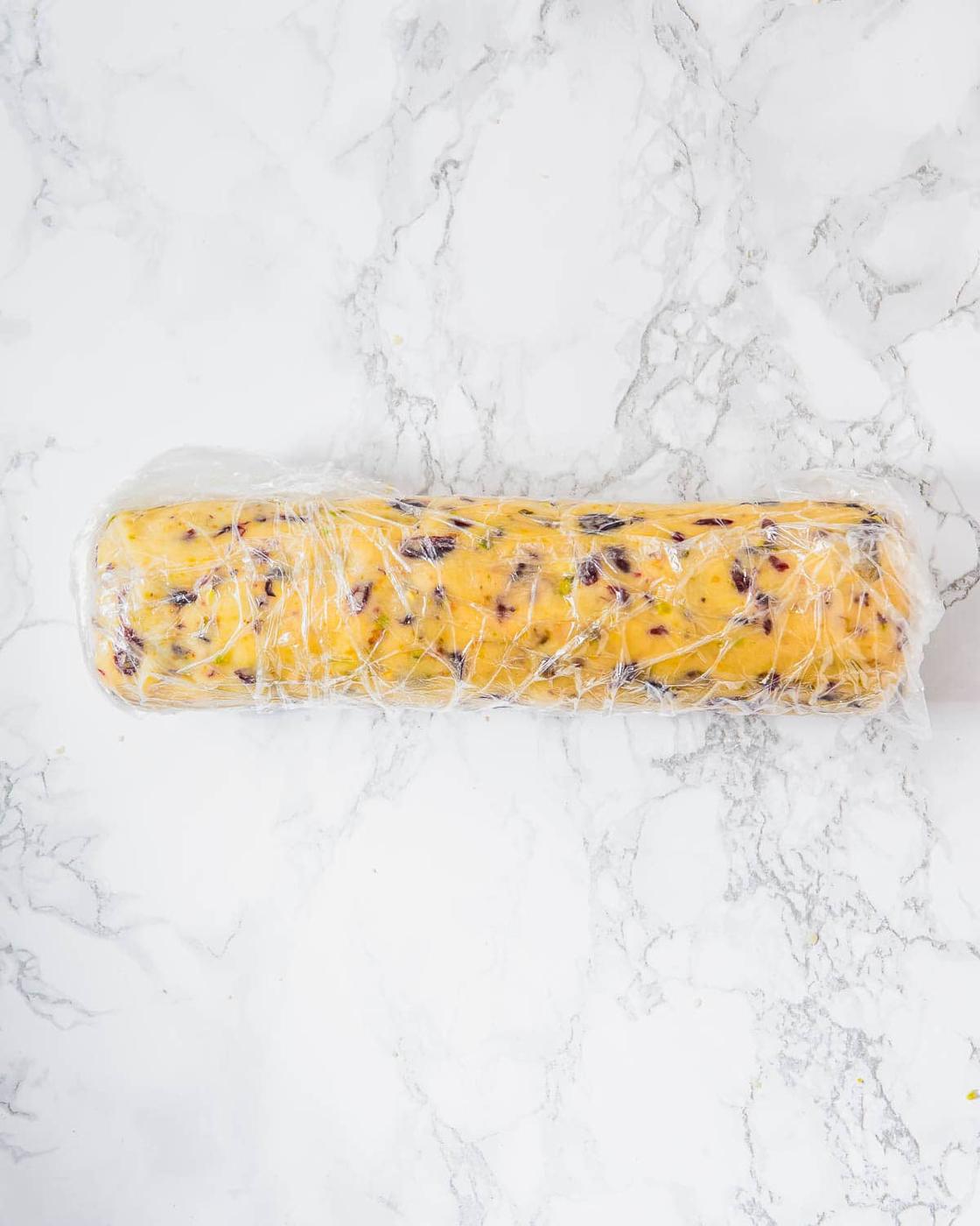 A log of shortbread dough with visible inclusions, wrapped in plastic wrap, resting on a white marble surface.