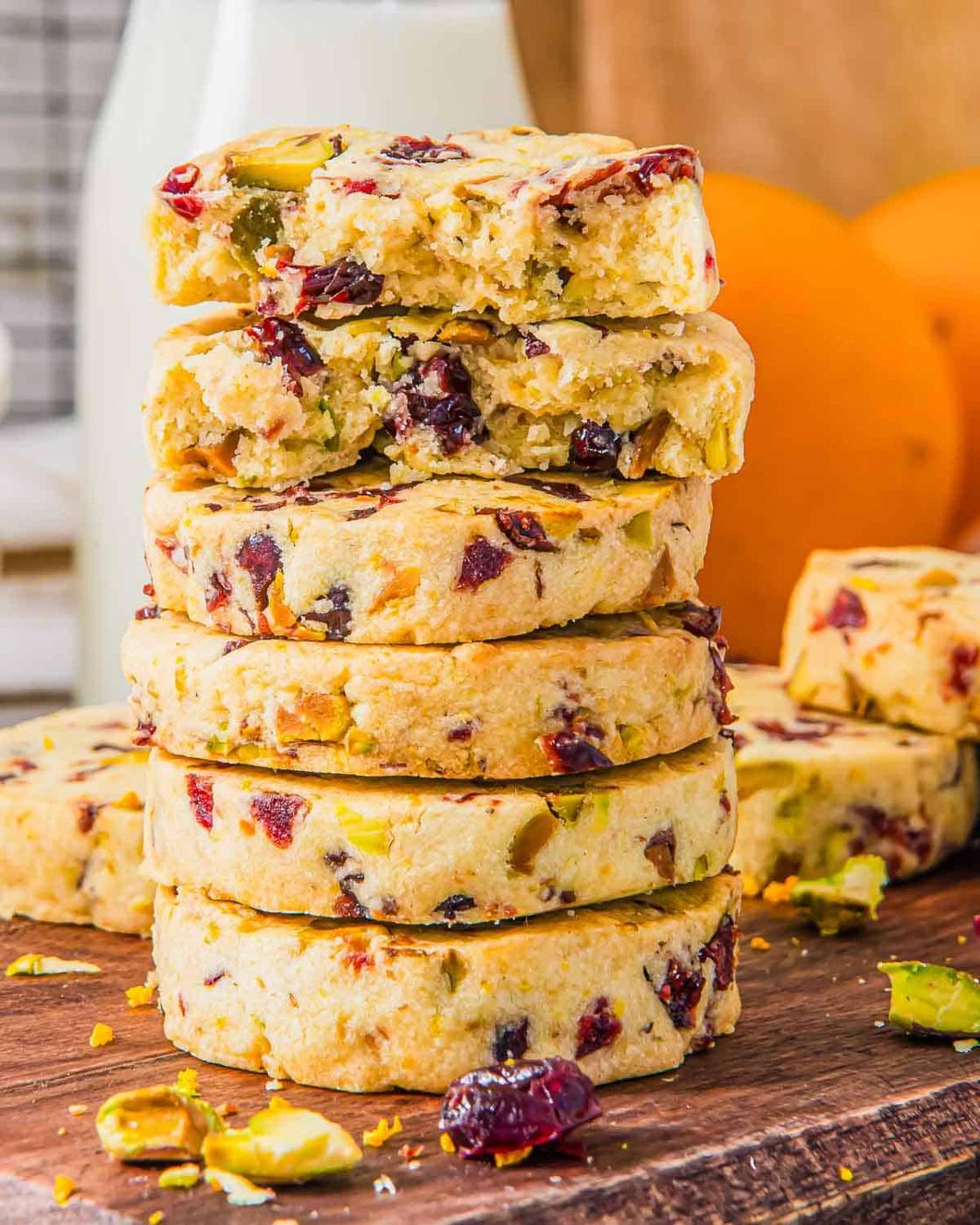 A stack of cranberry orange shortbread cookies on a wooden board. The cookies are studded with red cranberries and green pistachios. In the background, there's a bottle of milk and a whole orange.