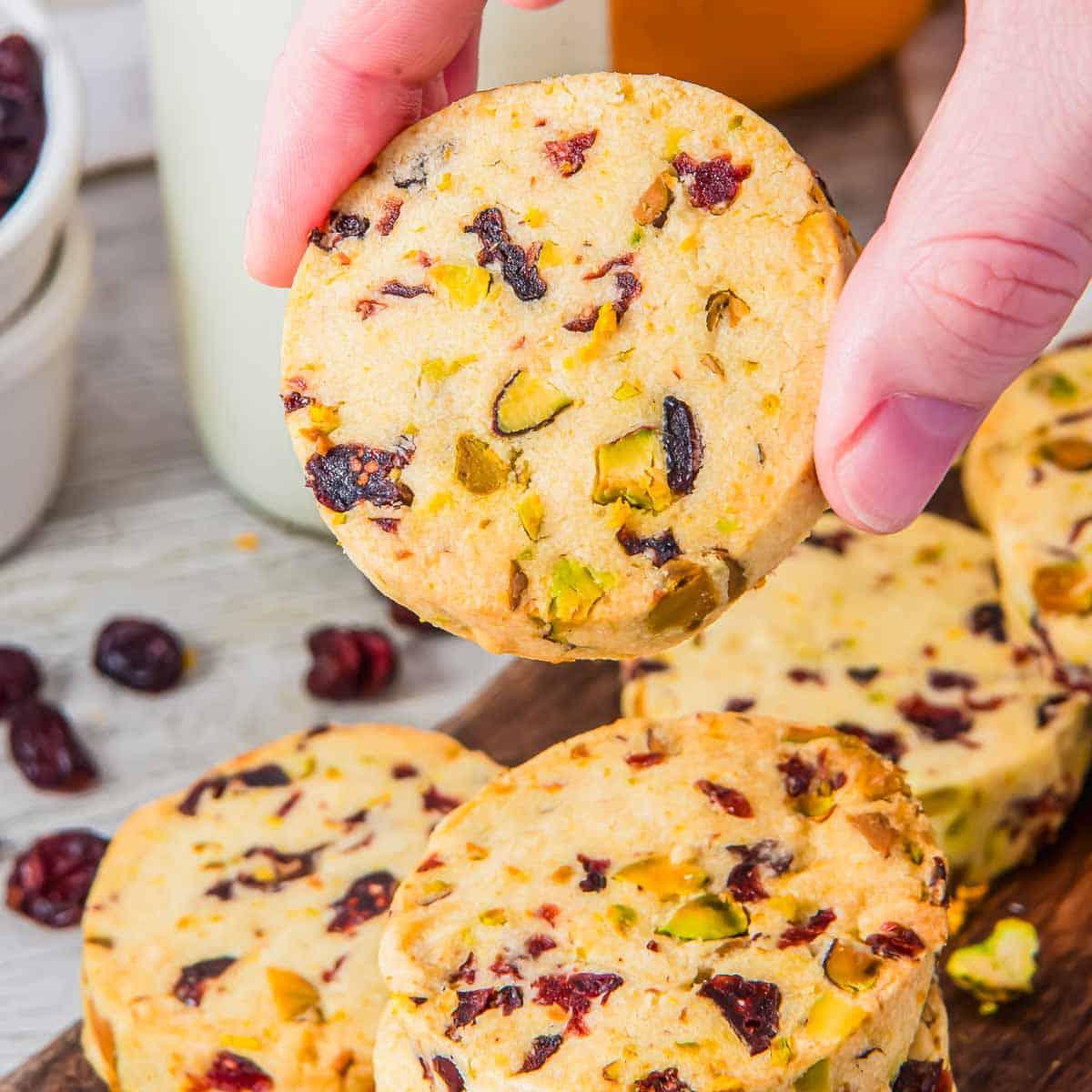 A hand holding a round shortbread cookie with visible chunks of orange zest, pistachios and cranberries. More cookies are stacked on a wooden surface below. A small bowl with cranberries and a glass of milk are in the background.