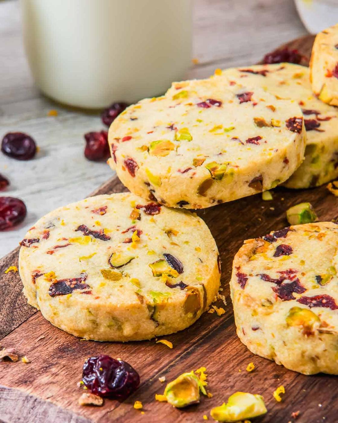 Shortbread cookies with pistachios and cranberries stacked on a wooden board. Cranberries, orange zest and scattered pistachios surround the cookies. A bottle of milk is visible in the background.