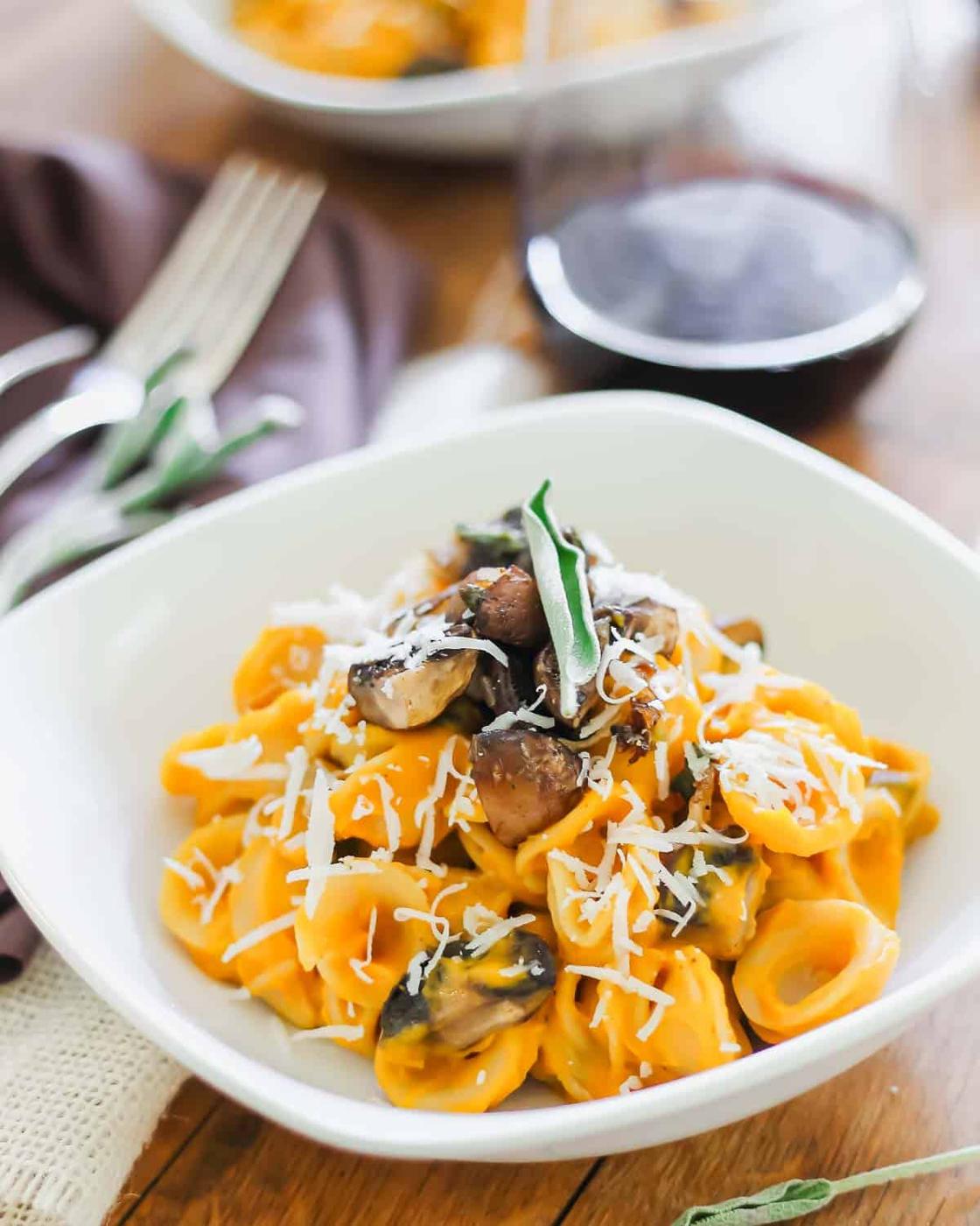 A bowl of creamy pasta topped with sautéed mushrooms, fresh sage, and grated cheese. A glass of red wine and a fork are placed beside the dish on a wooden table with a purple napkin.