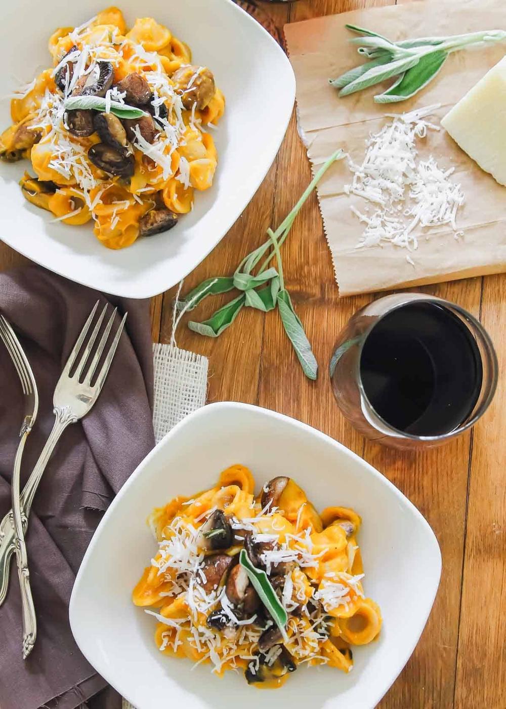 Two bowls of butternut squash pasta with mushrooms and garnished with shredded cheese and a sage leaf. A glass of red wine, cheese, sage sprigs, and utensils are on a wooden table.