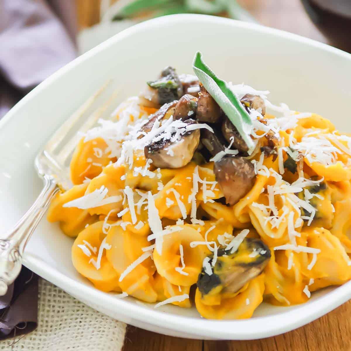 A bowl of pasta tossed with a creamy butternut squash sauce, mushrooms, and shredded cheese. A sprig of fresh sage garnishes the dish. A fork rests on the edge of the white bowl, which is set on a wooden table.