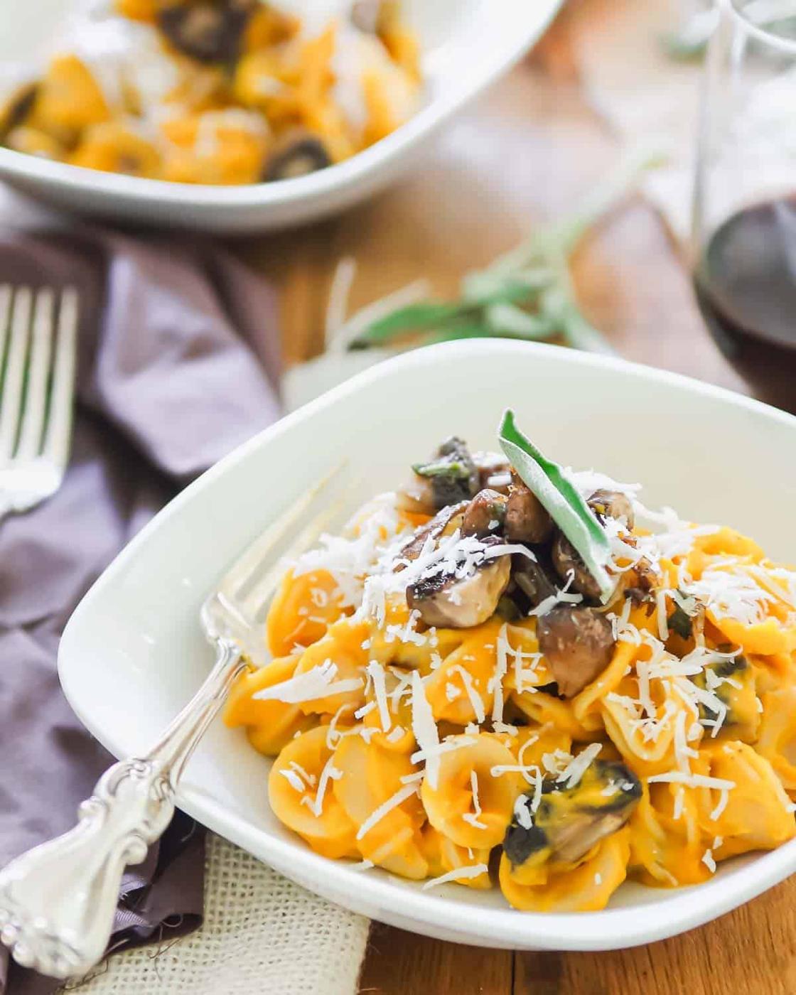 A white plate filled with creamy butternut pasta topped with grated cheese and mushrooms. A fork rests on the plate. A glass of red wine and another plate with tortellini are in the background. A napkin is placed beside the dish.