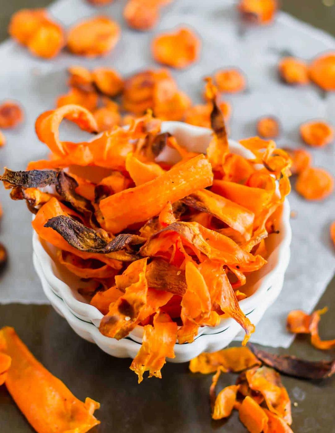 A bowl of homemade carrot chips on a gray surface, showcasing crispy, orange slices with slightly charred edges.