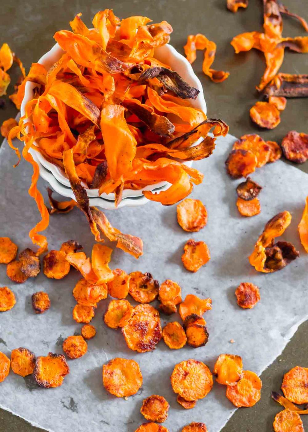 A bowl of crispy carrot chips atop a slate surface surrounded by additional scattered chips.