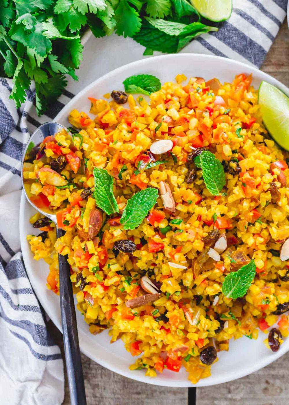 Frozen cauliflower rice with curry spices, raisins, almonds and mint garnish on a white plate.