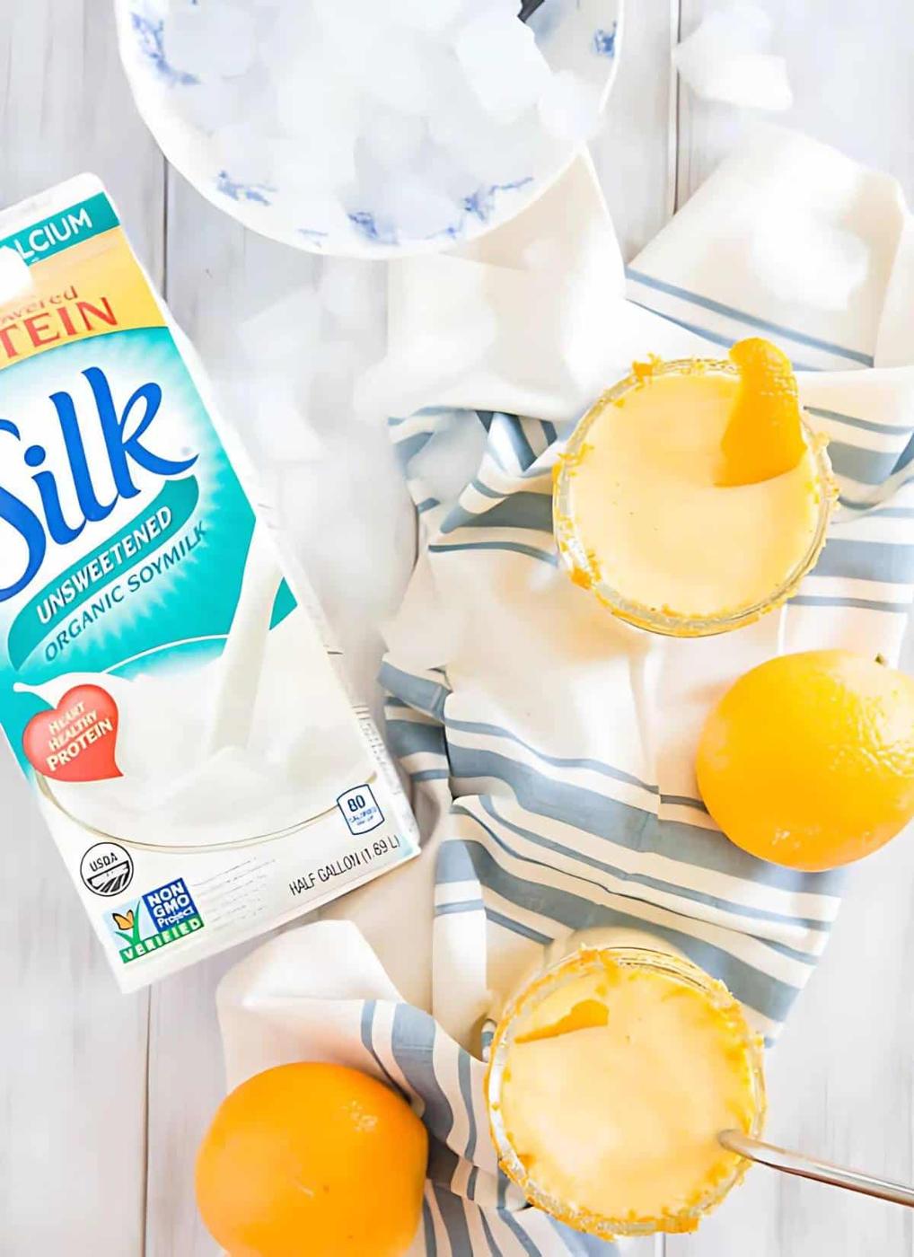 A carton of Silk unsweetened organic soy milk is beside two garnished glasses of a yellow beverage on a striped cloth, showcasing perfect ingredients for dairy-free cooking. Oranges and ice cubes are nearby.