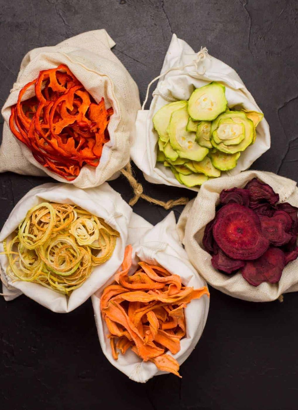 Five small cloth bags filled with colorful dried vegetable chips—orange carrot, green zucchini, yellow squash, purple beet, and orange sweet potato—arranged in a circle on a dark textured surface.
