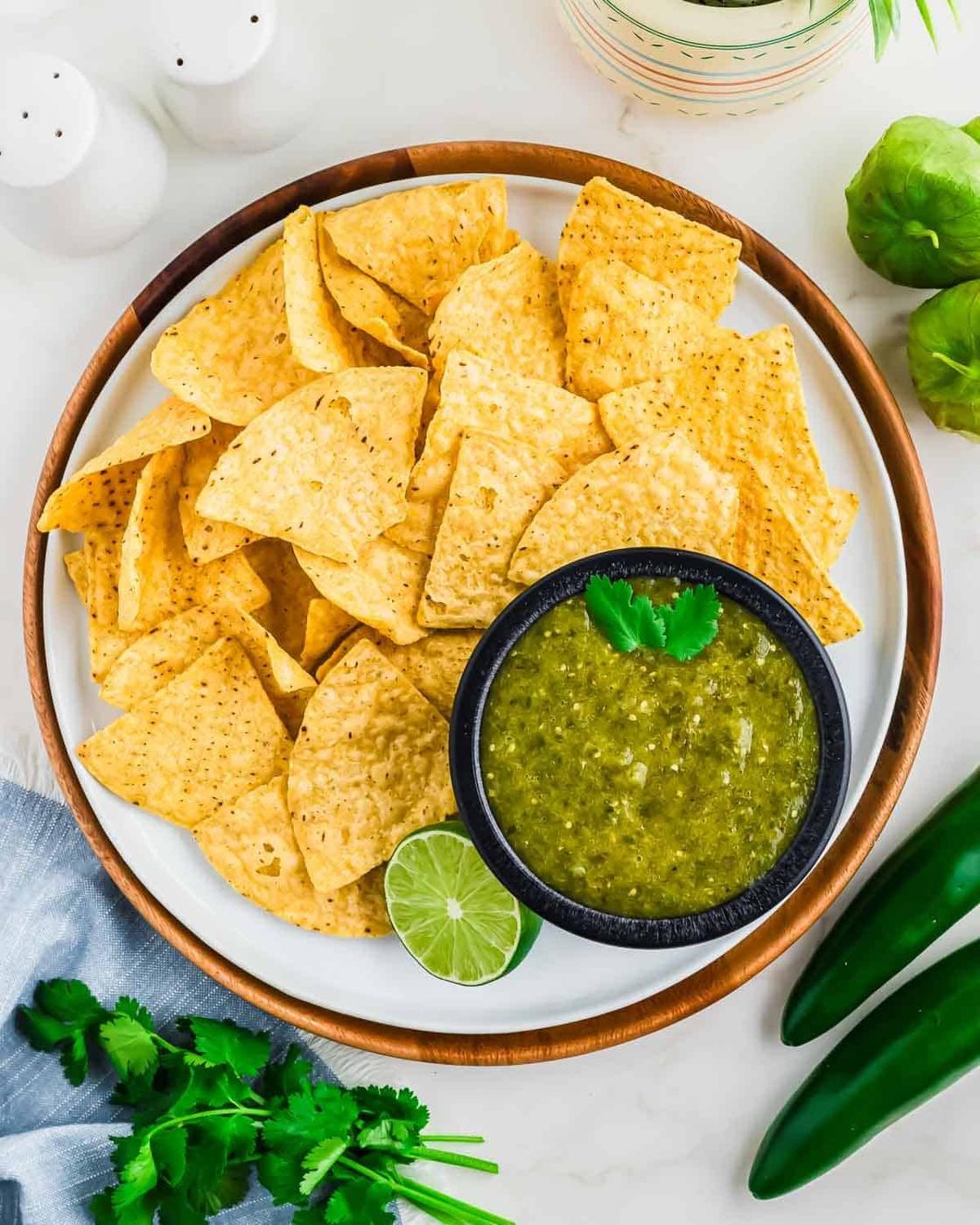 A plate of tortilla chips with a small black bowl of green salsa garnished with cilantro and a lime wedge, surrounded by fresh cilantro, jalapeños, and salt shakers on a white surface.