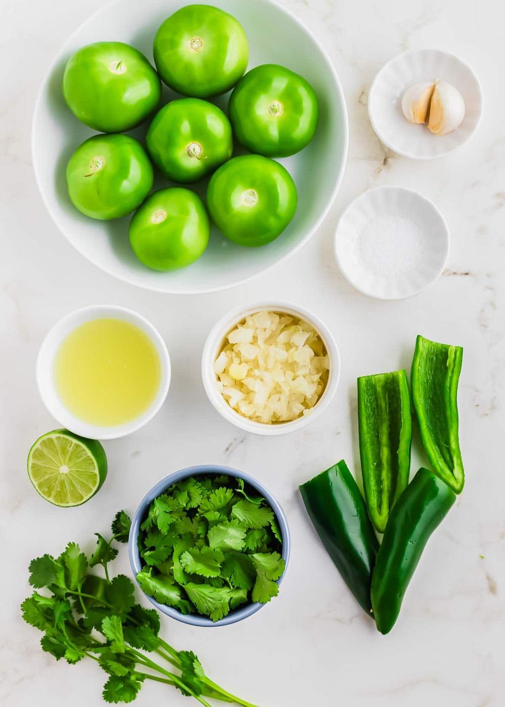 Overhead view of fresh ingredients on a white surface: whole tomatillos in a bowl, two garlic cloves, a bowl of salt, chopped onion, lime juice, halved jalapeños, cilantro, and half a lime.