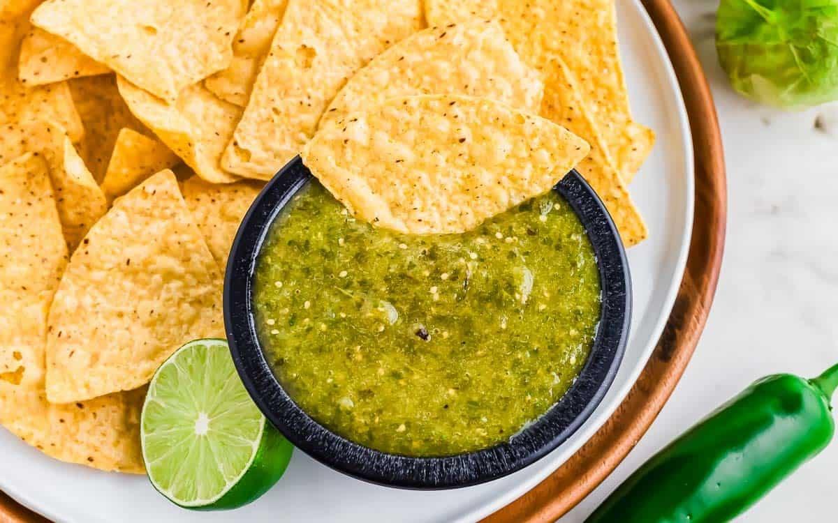 A bowl of homemade salsa verde with a tortilla chip dipped in it, surrounded by more tortilla chips on a plate. A halved lime and a jalapeño pepper are next to the bowl on a white surface.