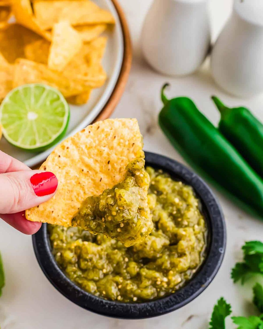 A hand with red nail polish dips a tortilla chip into a bowl of salsa verde. Nearby are fresh jalapeños, cilantro, a halved lime, and a plate of more chips.