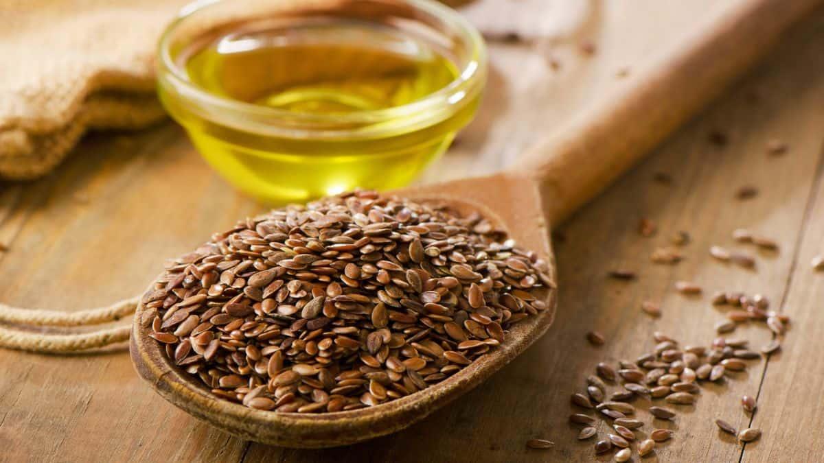 Flaxseeds on a wooden spoon with a bowl of flaxseed oil in the background.
