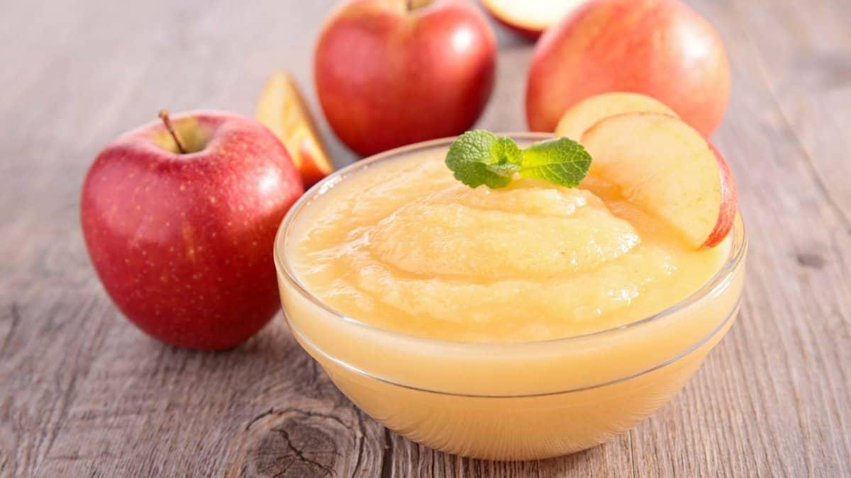 A glass bowl of applesauce garnished with apple slices and a mint leaf, placed on a wooden surface with whole red apples in the background.