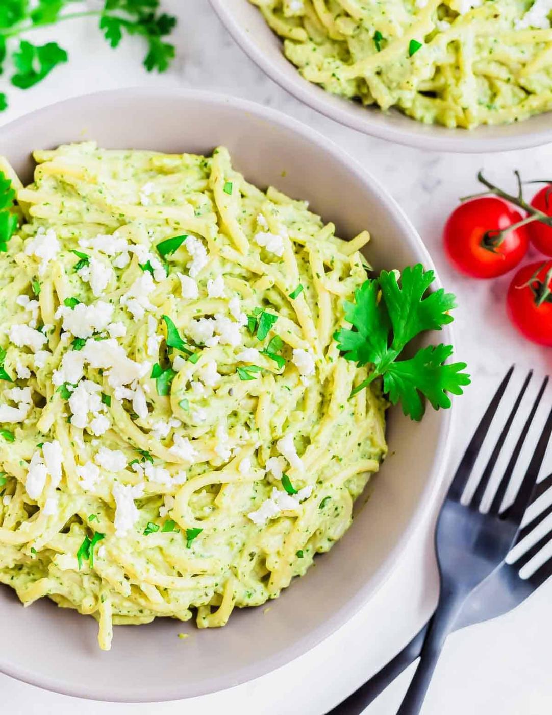 A bowl of creamy green pasta garnished with crumbled white cheese and fresh parsley, with a sprig of parsley and cluster of cherry tomatoes nearby. Black forks are placed beside the bowl on a white surface.