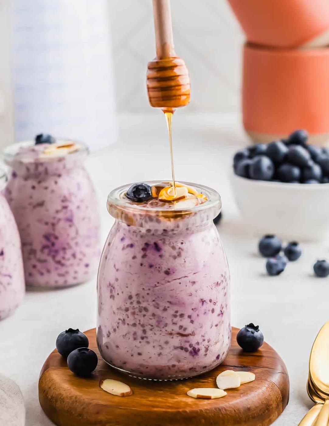 A jar filled with blueberry chia pudding topped with honey and blueberries. Other jars and bowls of blueberries are in the background.