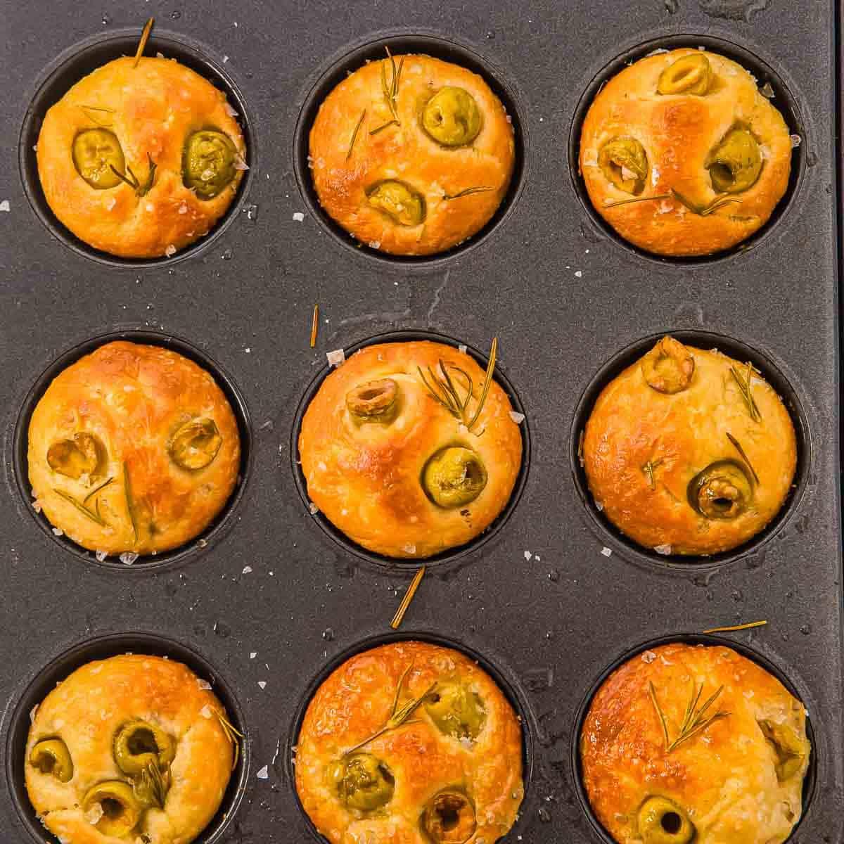 A muffin tin with nine baked focaccia muffin rolls topped with green olives, rosemary, and sea salt. The golden-brown rolls are evenly placed in each cup of the dark metal pan.