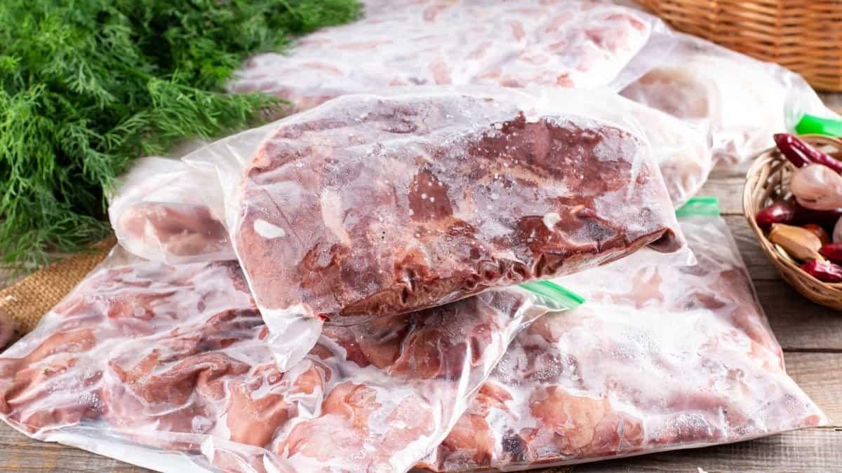 Several vacuum-sealed bags of frozen meat are stacked on a wooden surface. Fresh dill and a wicker basket with vegetables are partially visible in the background.
