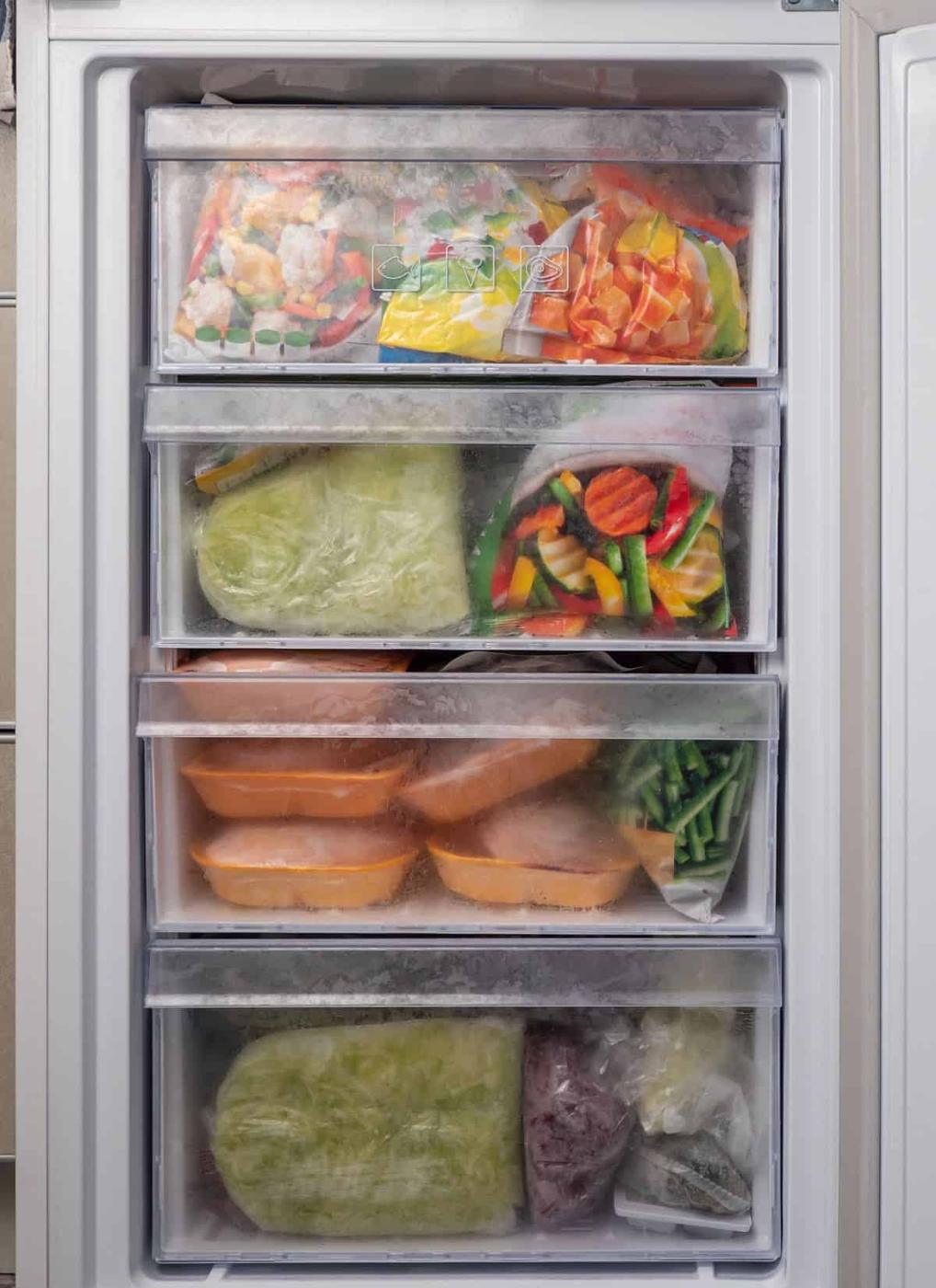 An open freezer with four drawers, each filled with various frozen foods, including vegetables, packaged meals, and plastic bags containing different items, all covered in a layer of frost.
